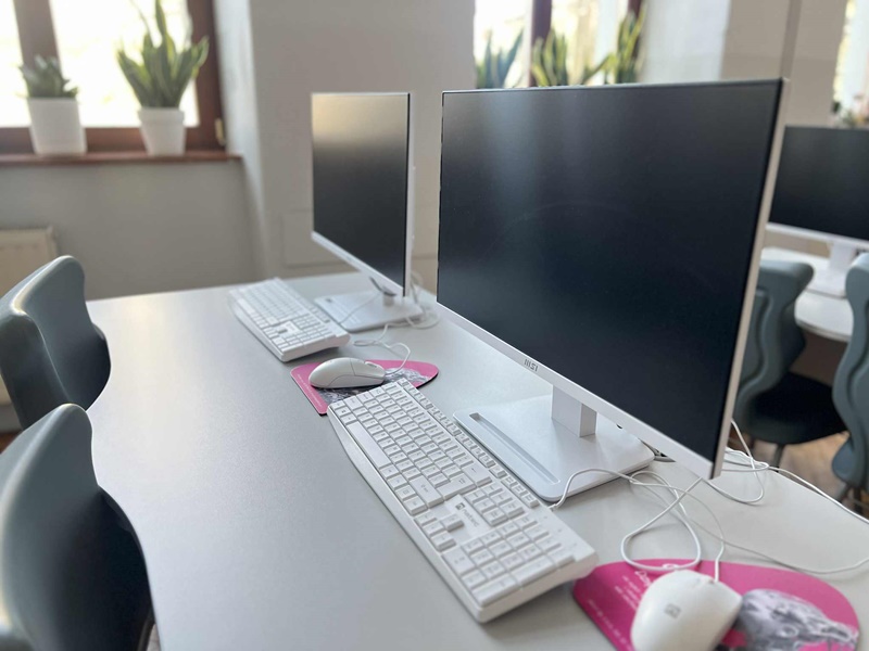 Rząd stanowisk komputerowych w jasnej sali: monitory, klawiatury i myszy ustawione na biurkach.